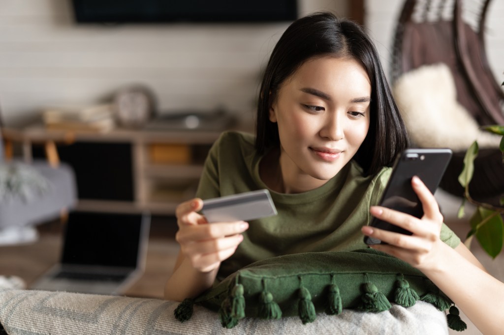 A young Asian woman who buys from the online store, using mobile phone and credit card, shopping from home.